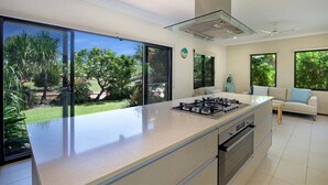 A modern kitchen with a large island, sliding glass doors to a garden, and a view of greenery. - Balu luxe (Broome)
