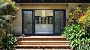 A modern house with a large sliding glass door, a wooden deck, and tropical plants. - Balu luxe (Broome)