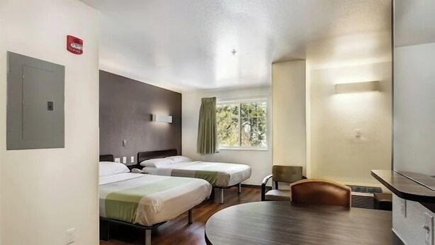 Ein Hotelzimmer mit zwei Einzelbetten, einem runden Esstisch, einem Stuhl, einem Fenster mit Vorhängen und einem an der Wand montierten Rauchmelder.