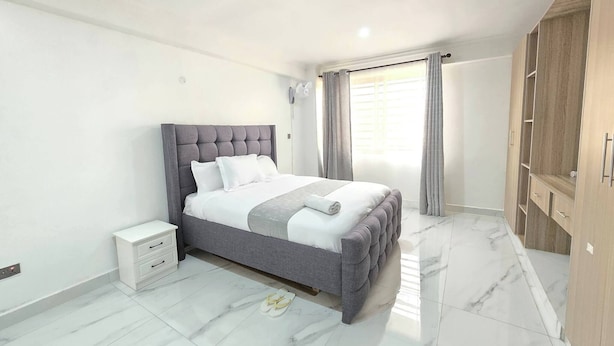 A bedroom with a bed, a nightstand, a wardrobe, and a window with curtains. - Serene 2-bedroom Apartment in Central Kisumu (Kisumu)