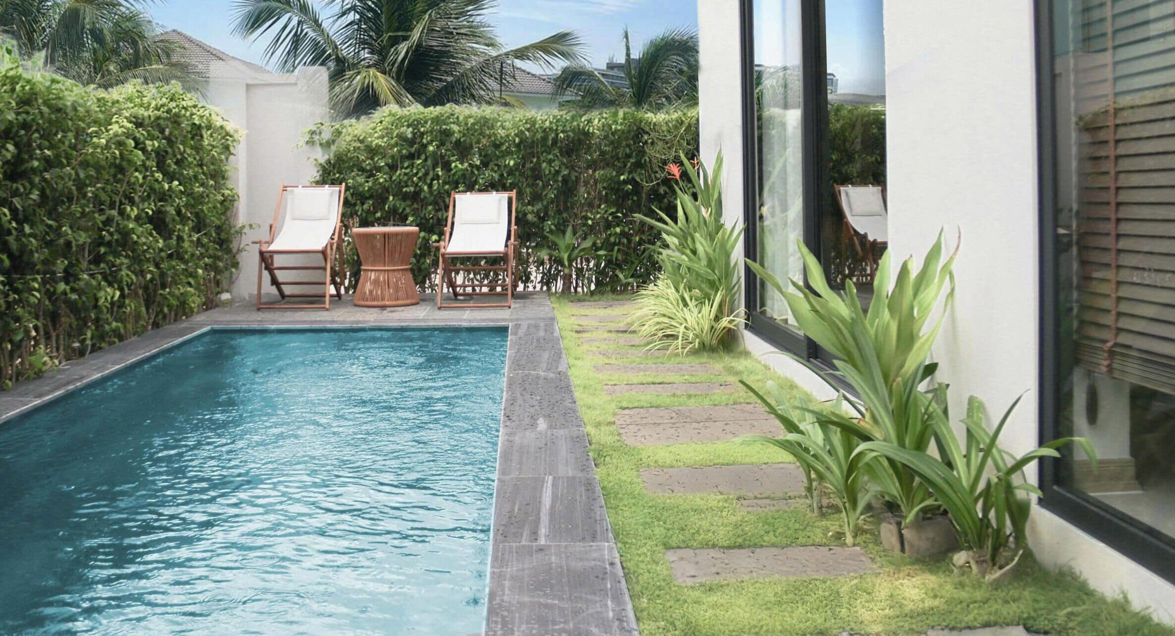 Una piscina con sillas, un pequeño jardín y un edificio con ventanas grandes.
