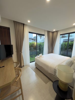 A modern hotel room with a bed, a desk with a chair, a TV, and a view of greenery outside. - Lagom Residence Phu Quoc (Phu Quoc)