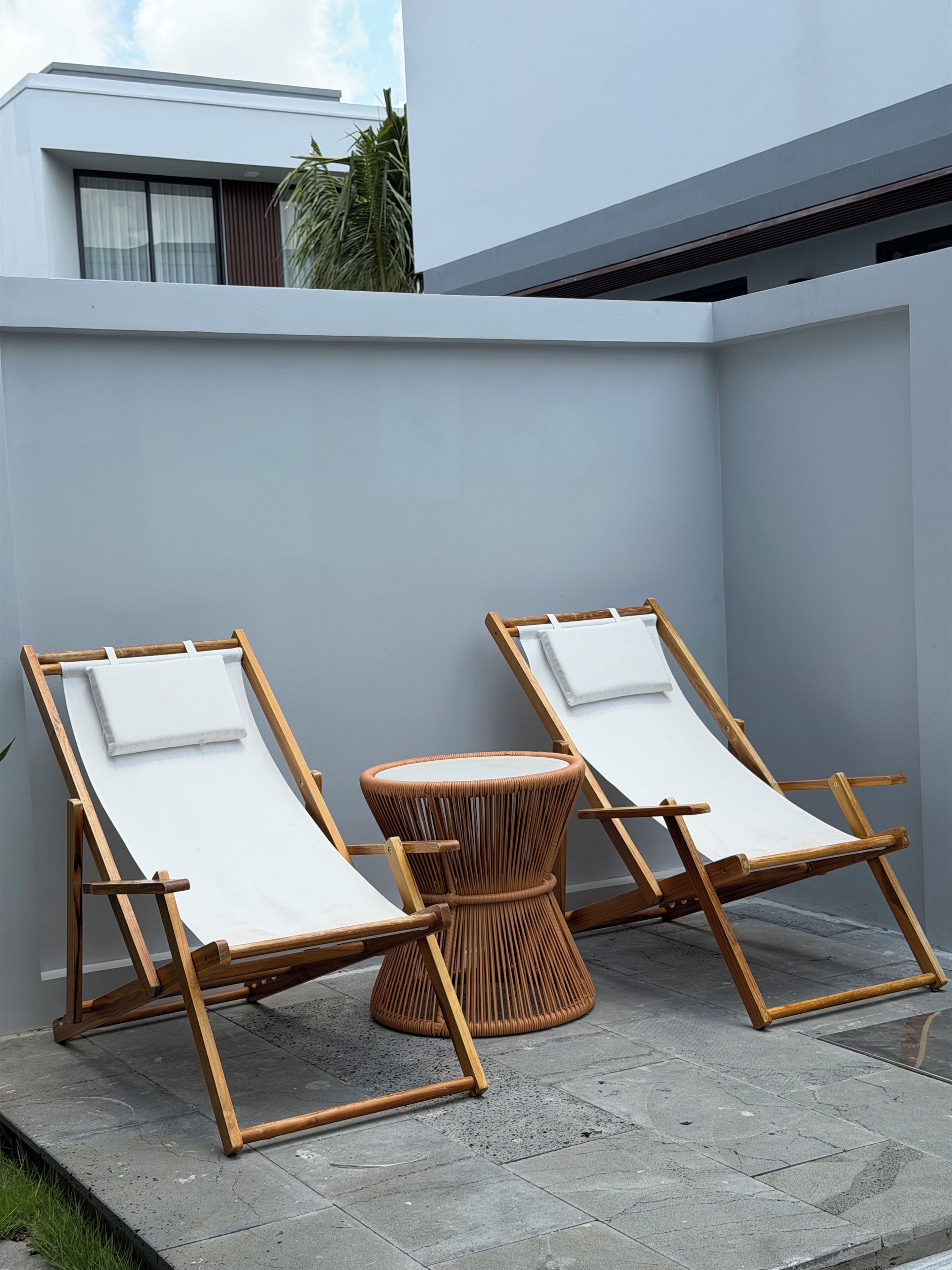 Dos sillones de madera con cojines blancos, una mesa redonda tejida y una zona de patio con suelo de baldosas grises.