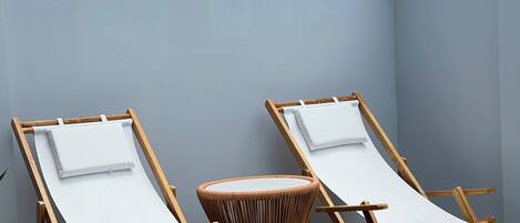 Dos sillones de madera con cojines blancos, una mesa redonda tejida y una zona de patio con suelo de baldosas grises.