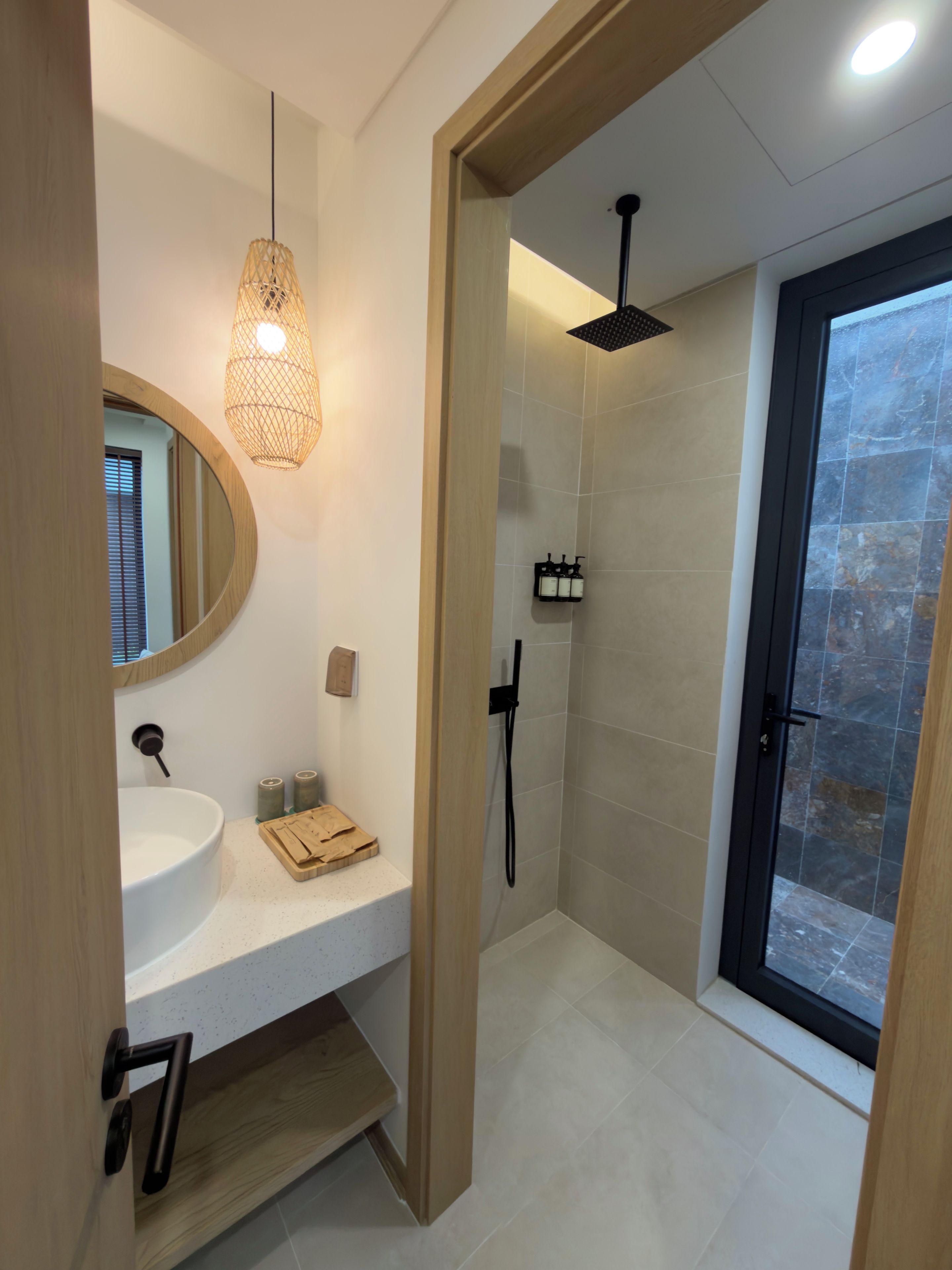 A modern bathroom with a round mirror, a white sink, a wooden shelf, a shower area, and a glass door.