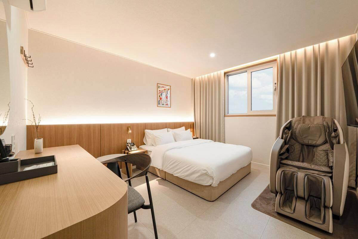 Ein Hotelzimmer mit Bett, Schreibtisch, Stuhl, Massagesessel und einem Fenster mit Vorhängen.