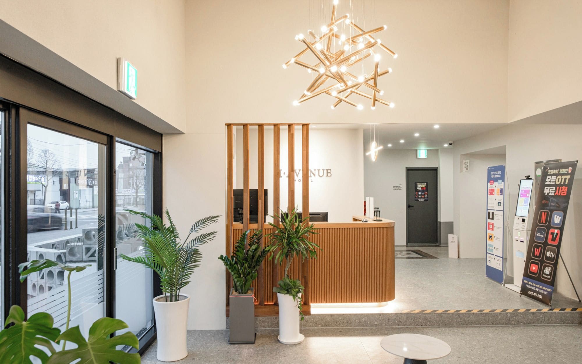 A hotel reception area with a wooden reception desk, potted plants, a modern light fixture, and a digital information kiosk.