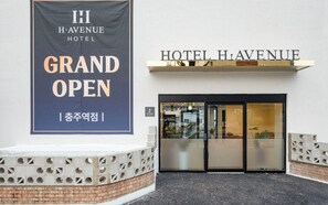 L’entrée de l’hôtel HI HAVENUE est marquée par une banderole sur laquelle on peut lire « GRANDE OUVERTURE » et une enseigne au-dessus de la porte indiquant « HÔTEL HAVENUE ».