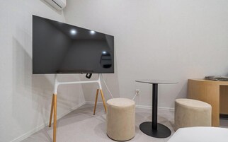 Una habitación moderna con un televisor de pantalla plana sobre un soporte blanco, una mesita redonda y dos otomanas.