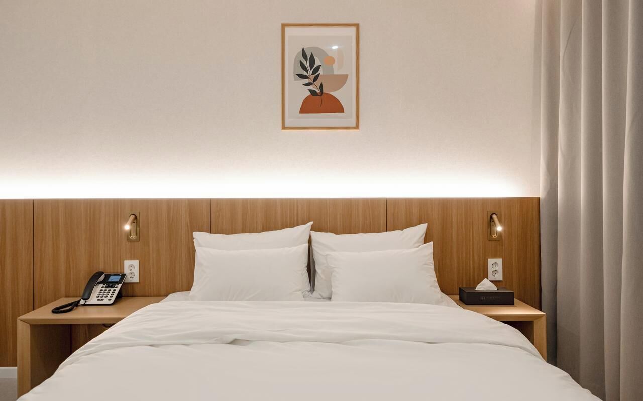 Ein Hotelzimmer mit einem Holzkopfteil, einem Bett mit weißer Bettwäsche, einem Nachttischtelefon und einem gerahmten Kunstwerk an der Wand.