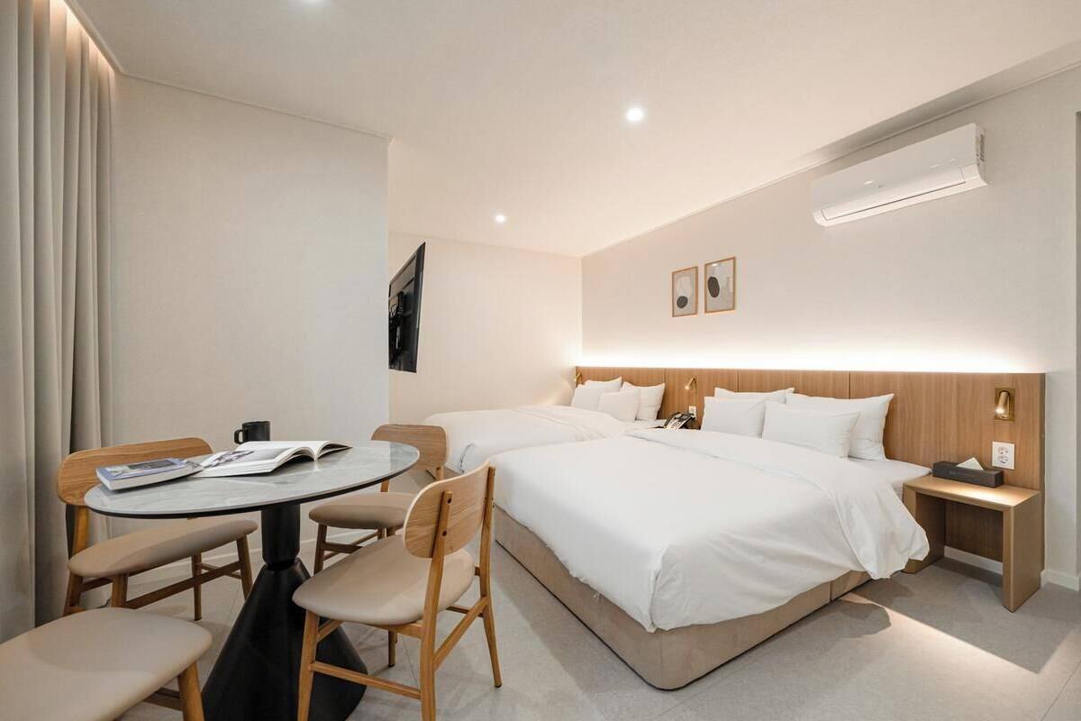 Ein Hotelzimmer mit zwei Betten, einem runden Tisch mit Stühlen, einem Nachttisch mit Lampe und einer Klimaanlage.