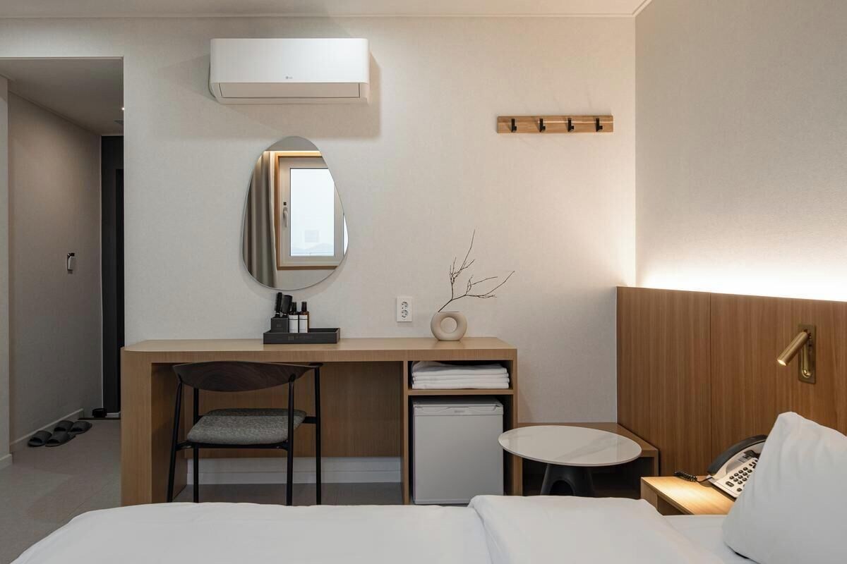 Una camera d'albergo con un letto, una scrivania, una sedia, un tavolino, uno specchio, un telefono, un termosifone e un appendiabiti.