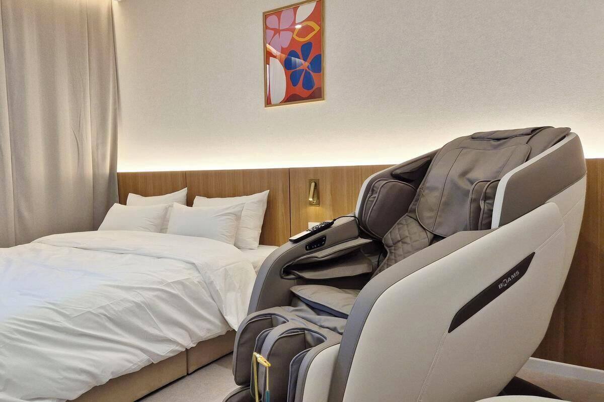 Ein Hotelzimmer mit Bett, Massagesessel, Massageliege an der Wand und einem Gemälde.