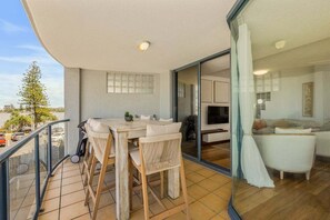 A balcony with a table and chairs, a sliding glass door leading to a living area with a sofa and TV. - 4/16 Leeding Terrace, Caloundra QLD 4551 (Sunshine Coast)
