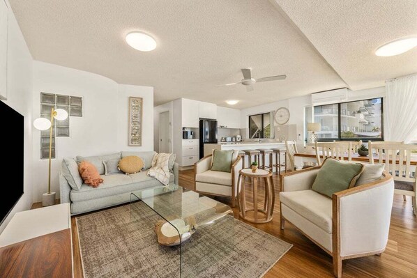 A modern living room with a glass coffee table, a sofa, and two armchairs. There is a kitchen area with a refrigerator and a dining table with chairs. - 4/16 Leeding Terrace, Caloundra QLD 4551 (Sunshine Coast)