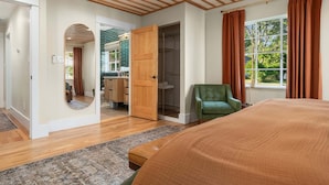 Ein Schlafzimmer mit einem großen Bett, Holzboden, einem grünen Sessel, einem begehbaren Kleiderschrank und einem Fenster mit Vorhängen.