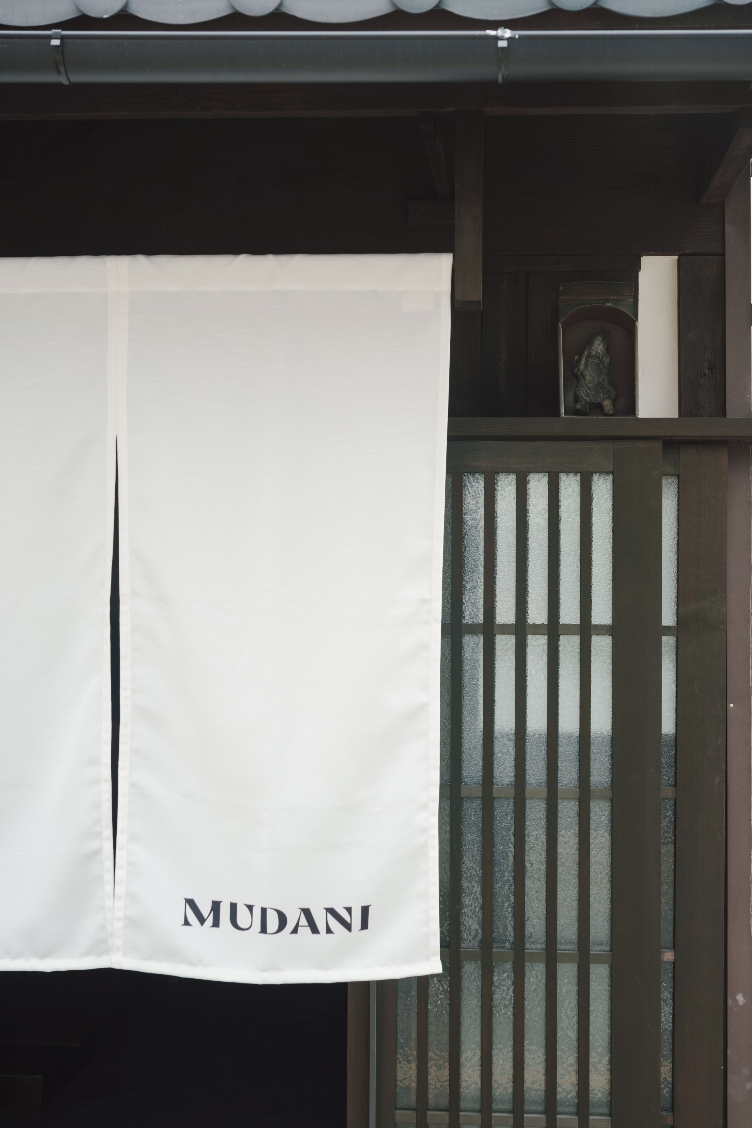 A white banner with 'MUDANI' text hanging in front of a wooden gate with vertical slats.