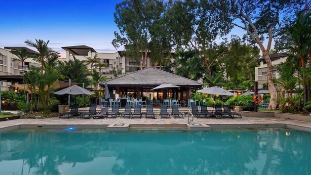 A swimming pool with lounge chairs and umbrellas, surrounded by trees and buildings.