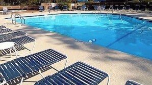 Una piscina con sillones de descanso, un edificio de madera y árboles al fondo.