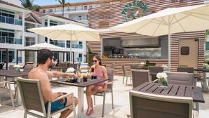 Un área de comedor al aire libre con mesas y sillas, un bar y un edificio con fachada a rayas.