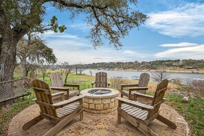 A circular stone fire pit with four wooden Adirondack chairs around it, overlooking a lake and distant buildings.