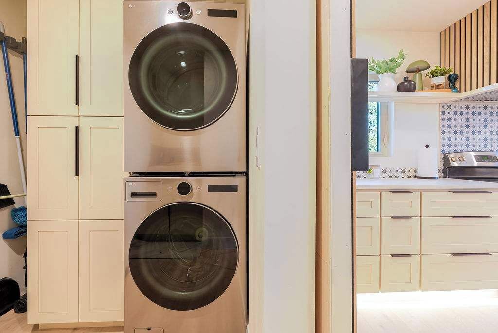 A laundry room with two stacked washing machines, a kitchen with white cabinets, and a window with patterned curtains.