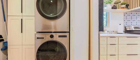 A laundry room with two stacked washing machines, a kitchen with white cabinets, and a window with patterned curtains.