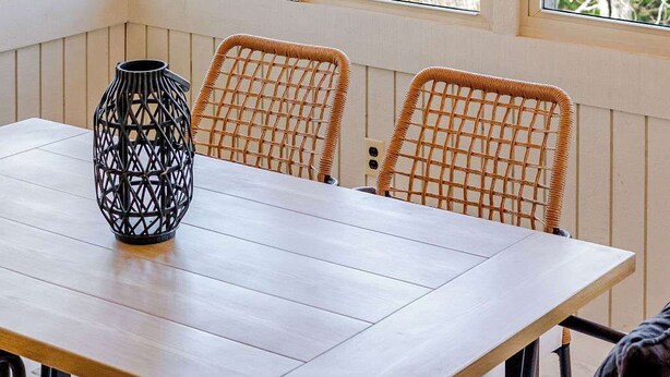 Ein Essbereich mit einem Holztisch, Korbstühlen, einer schwarzen Vase und mehreren Fenstern mit Tageslicht.