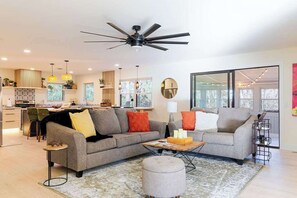 A modern living room with a sofa, ottoman, and coffee table, a ceiling fan, and a kitchen in the background.
