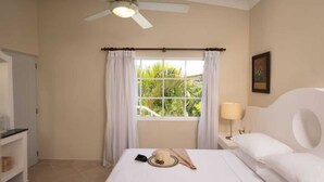 A hotel room with a bed, a window with curtains, a ceiling fan, a bedside lamp, and a painting on the wall.