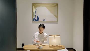 Uma pessoa vestindo uniforme branco preparando comida em uma mesa de madeira, com uma luminária e uma obra de arte emoldurada acima.
