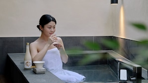 Eine Person in einer Badewanne mit Handtuch, Flasche und Tasse auf einer Ablagefläche.