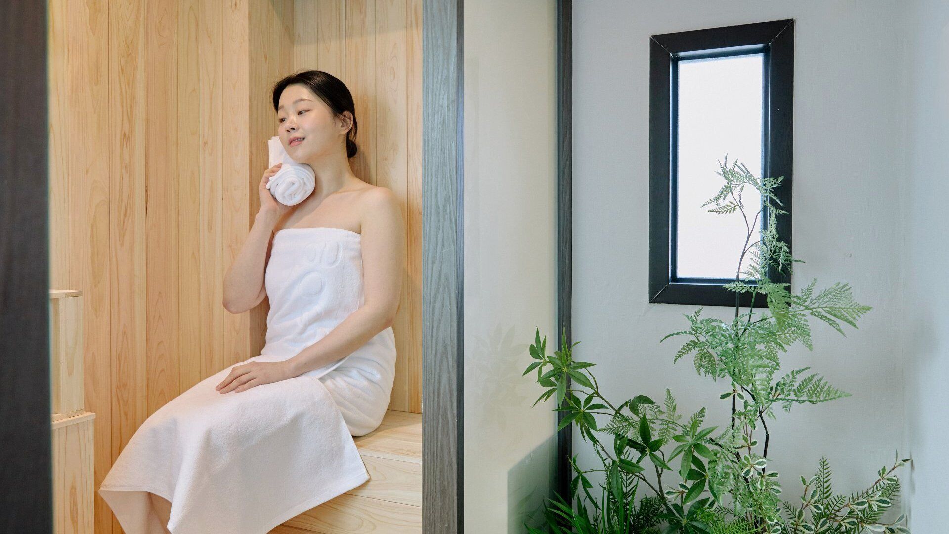 Una persona sentada en una habitación de madera, envuelta en una toalla blanca, con un espejo y plantas.
