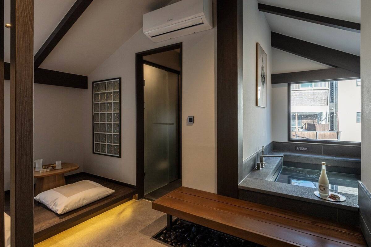 A hotel room with a bed, a small table with a bottle, a bench, a window with a view, and an air conditioning unit.