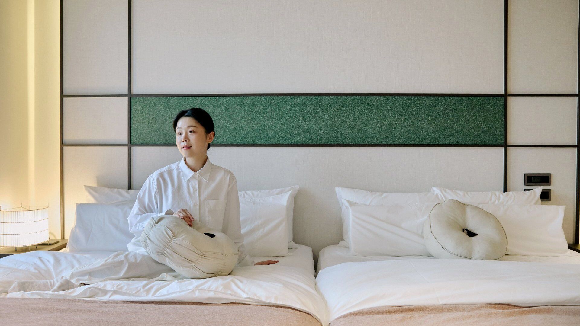 A hotel room with a bed, pillows, a bedside table, and a wall with a green panel.
