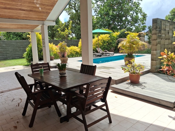 A covered patio with a pool, wooden table and chairs, potted plants, and a stone wall.