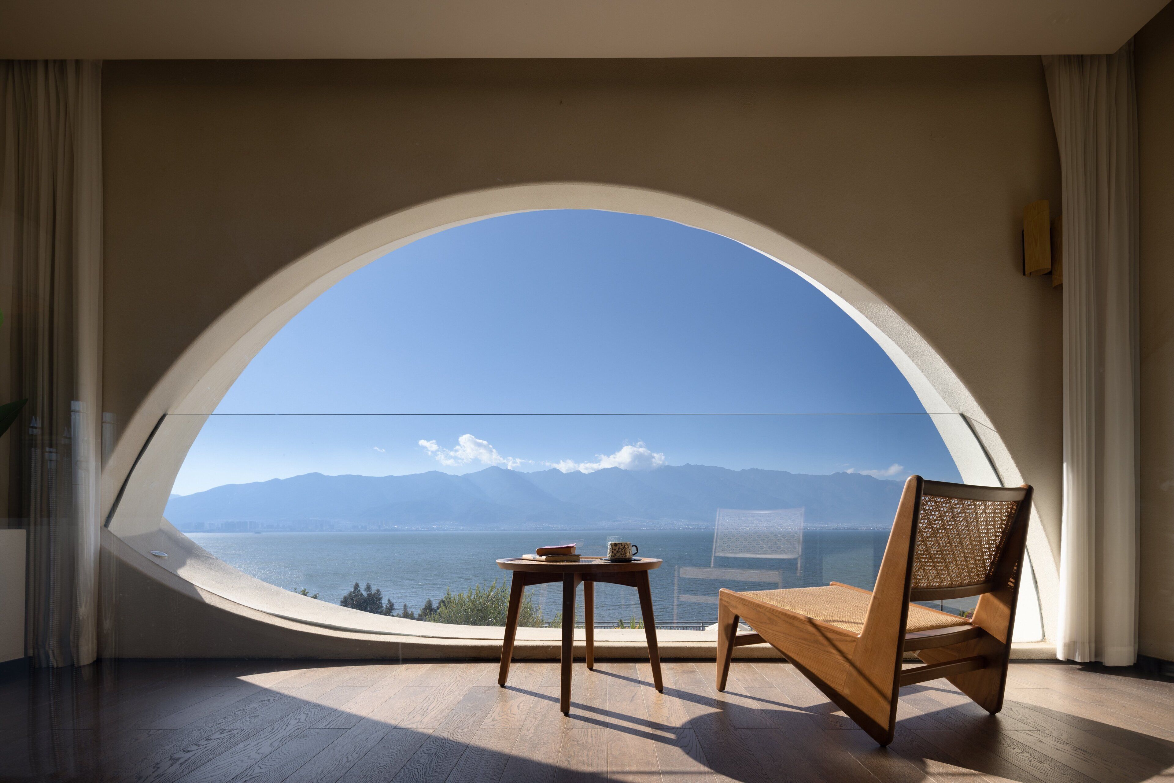 A room with a large arched window overlooking a body of water and mountains, a wooden chair, and a small table with a book and a mug.