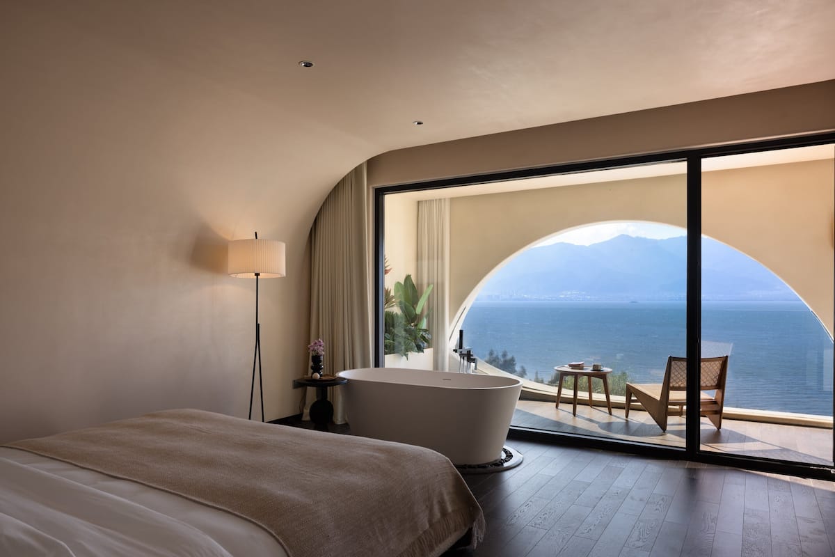 A bedroom with a large bed, a bathtub, a small table, and a chair, with a view of the sea and mountains through a large window.