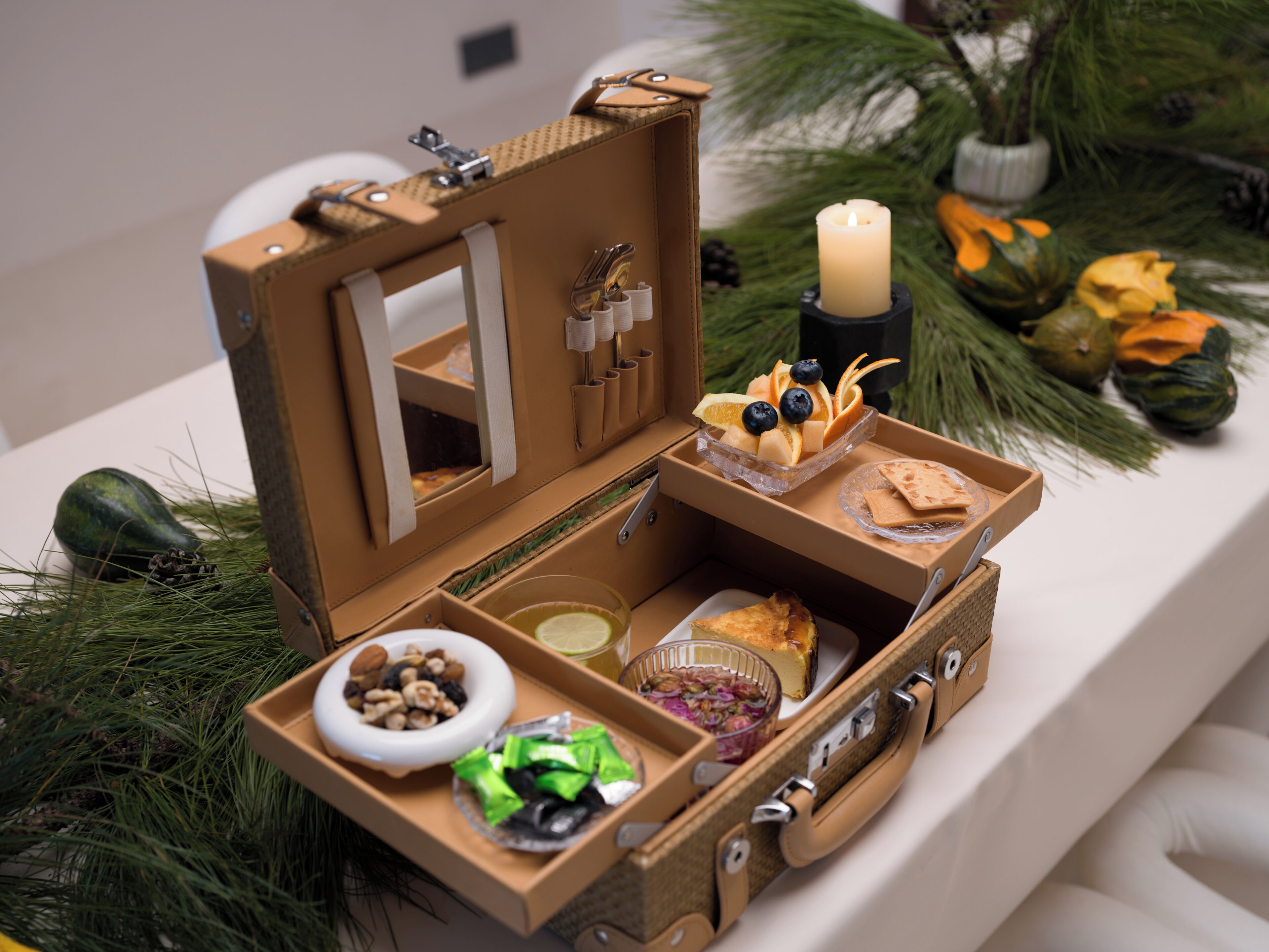 A small, open suitcase with compartments containing food items, a candle, and decorative pine branches.