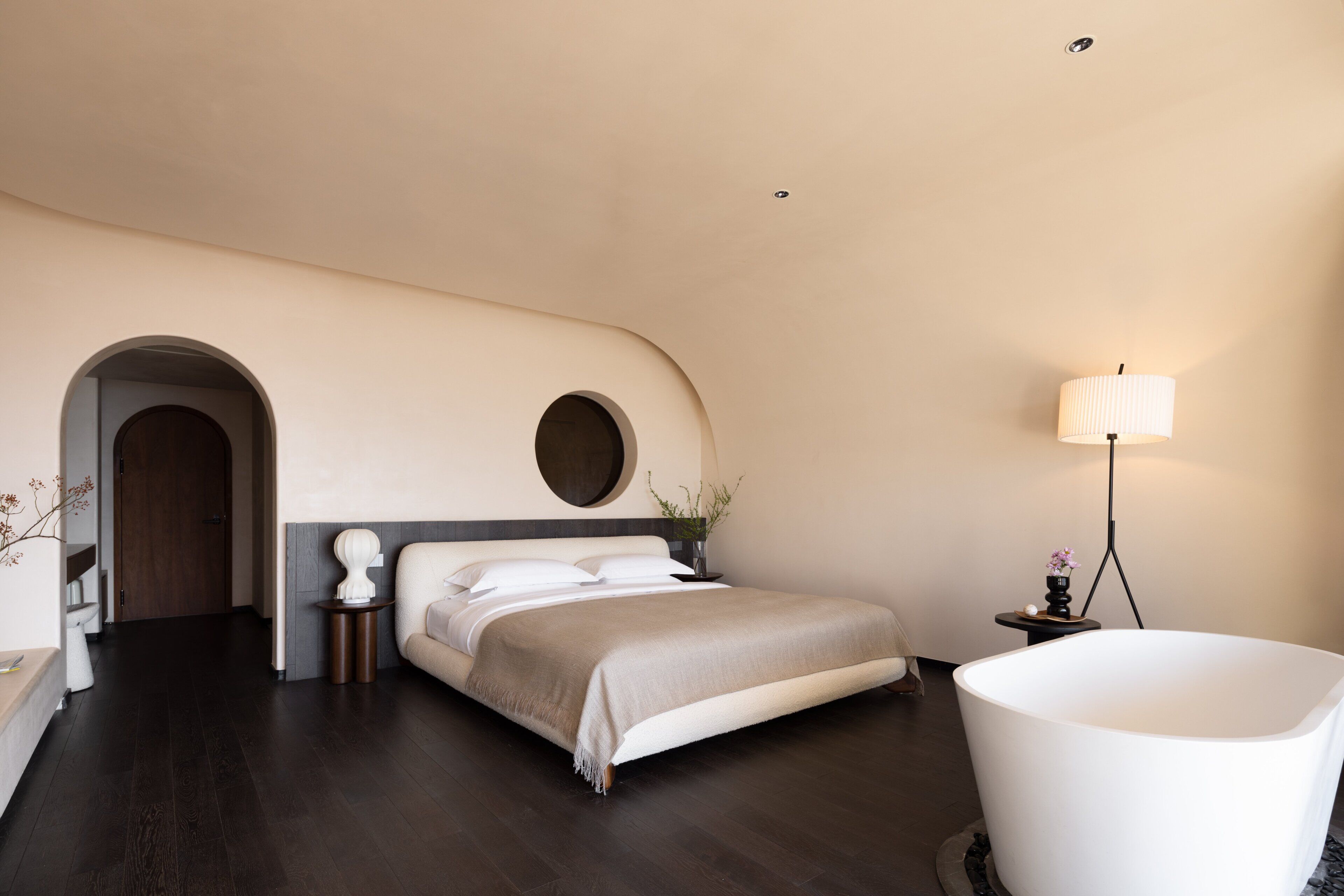 A modern bedroom with a large bed, a round mirror, a bedside table with a lamp, and a freestanding bathtub.