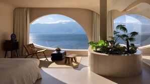 Une pièce comprenant un lit, une chaise, une petite table, une plante en pot et une vue sur la mer, visible à travers des fenêtres en arc de cercle.