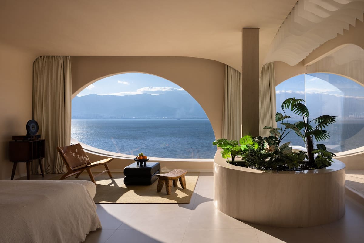 A room with a bed, a chair, a small table, a potted plant, and a view of the sea through arched windows.
