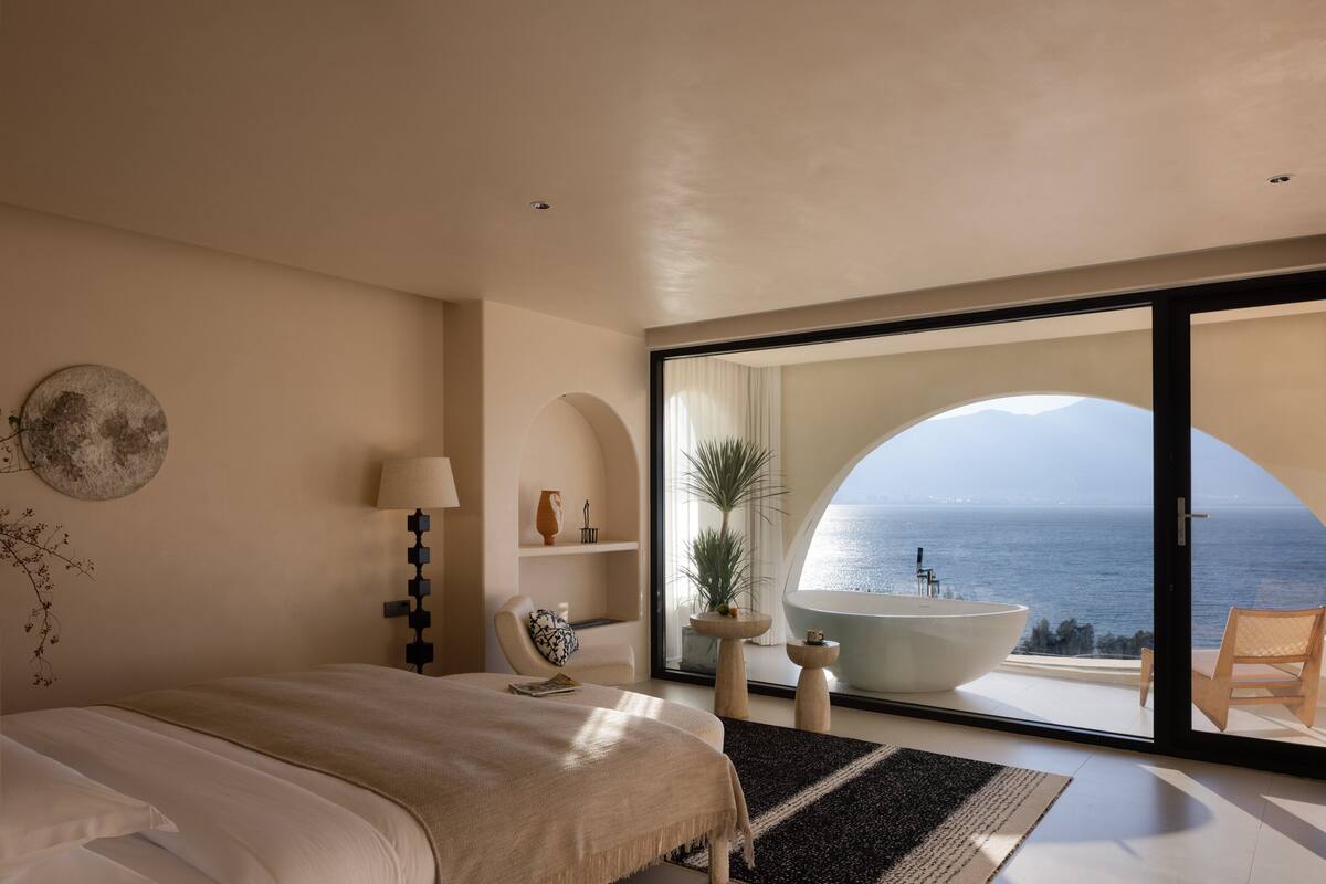 A bedroom with a large bed, a view of the sea through a glass door, a bathtub, and a chair by the window.