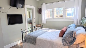 Une chambre à coucher comprenant un lit, un téléviseur fixé au mur, une fenêtre avec des rideaux et une porte donnant sur une salle de bain.