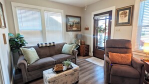 Un salon avec un canapé marron, un fauteuil assorti, un pouf à motifs et un ventilateur de plafond.