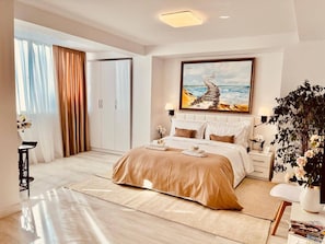 A modern bedroom with a large bed, a painting of a coastal scene, a potted plant, and a view of a cityscape through the window. - Leo s Garden View Apartment Cozy 1BR (Tirana)