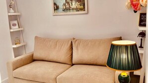 A living room with a beige sofa, a framed painting, a small side table with a lamp, and a vase with flowers. - Leo s Garden View Apartment Cozy 1BR (Tirana)