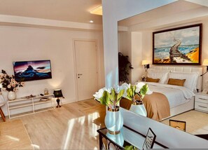 A modern hotel room with a bed, a TV, a desk, and a painting of a coastal scene. - Leo s Garden View Apartment Cozy 1BR (Tirana)
