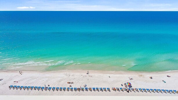 Una playa con hileras de sillas azules y sombrillas, una orilla arenosa y un océano azul cristalino.