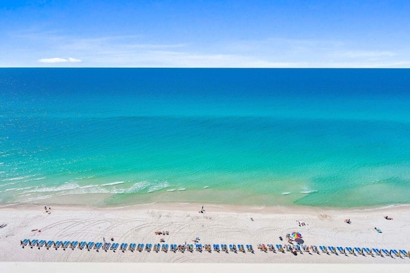 A beach with rows of blue lounge chairs and umbrellas, a sandy shore, and a clear blue ocean. - 🌴 New Luxury Gulf-Front Escape | Fully Remodeled with Stunning Views 🌊 (Panama City Beach)